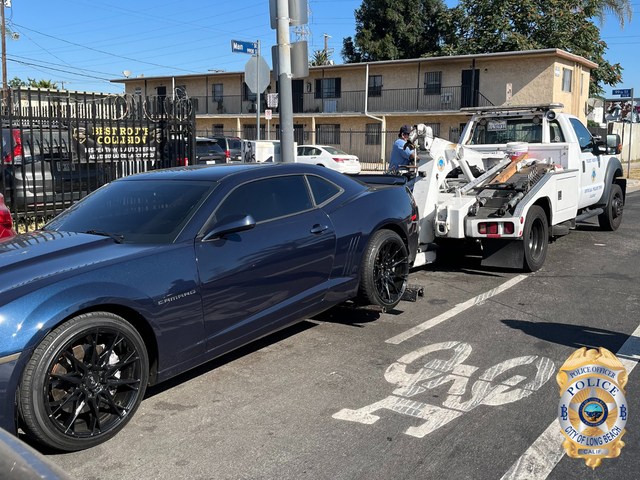 A dark colored car is hooked up to a white tow truck next to an apartment building.