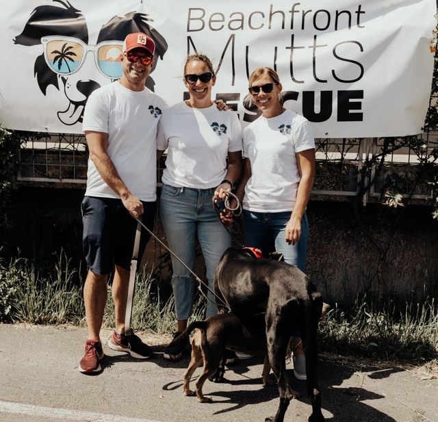 Three people and two dogs stand in front of a sign saying "Beachfront Mutts Rescue."