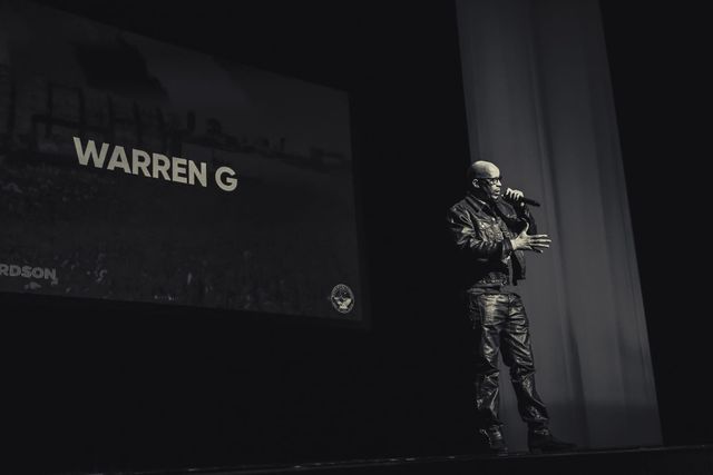 A man on stage with a microphone stands in front of a screen saying "Warren G."