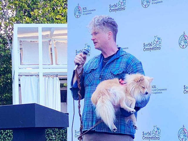 A man stands at a lectern holding a microphone and a small beige dog.