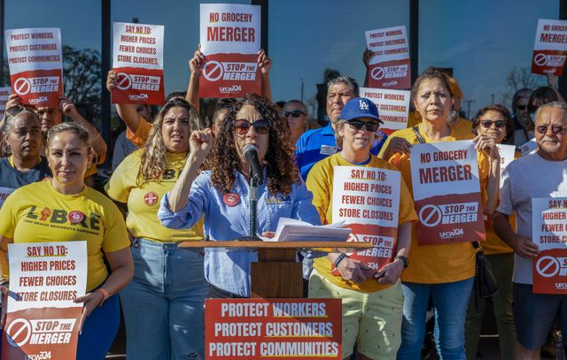 A woman speaks at a microphone in front of many people holding signs saying "Stop the merger."