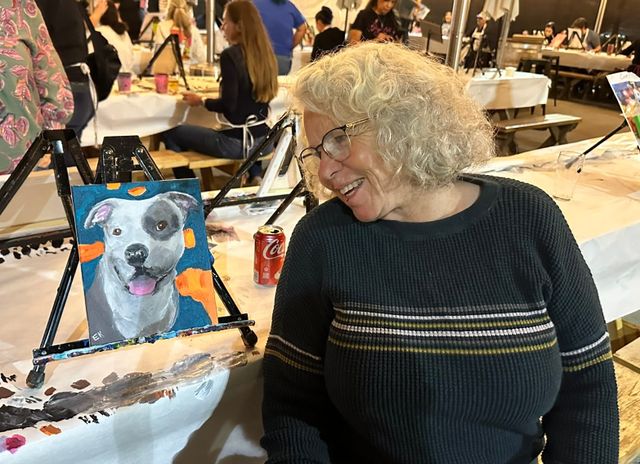 A woman in a black sweater smiles as she looks at a portrait of a white dog with a gray circle surrounding one eye.