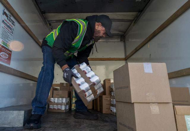 A man in a reflective vest loads boxes of canned food into a truck.