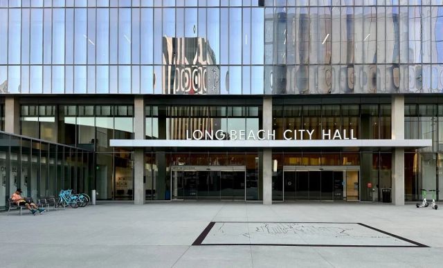 A man relaxes in a chair next to a glassy office building bearing a sign saying "Long Beach City Hall."