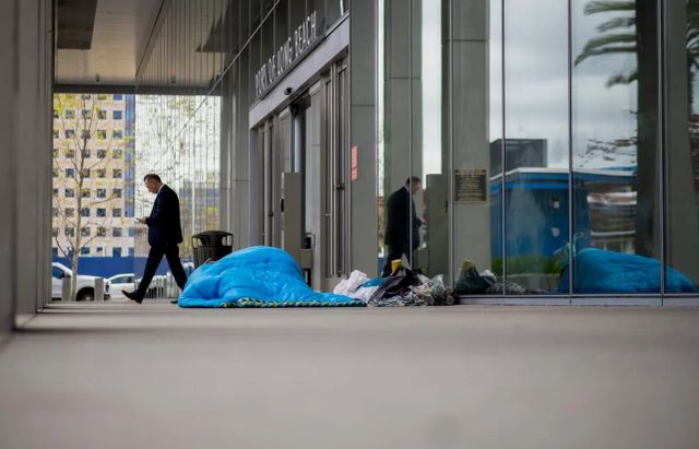 A person lays beneath a bright blue blanket next to the front door of a building labeled "Port of Long Beach" as a man in a suit walks by.