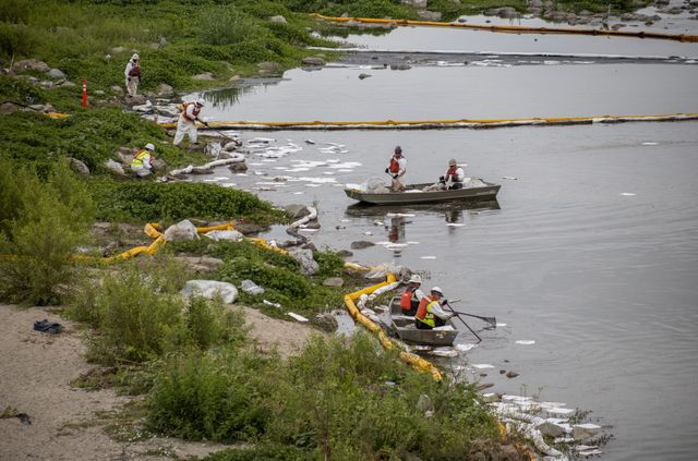 People in white coveralls and hats stand near a very dirty riverbank and in small gray boats.