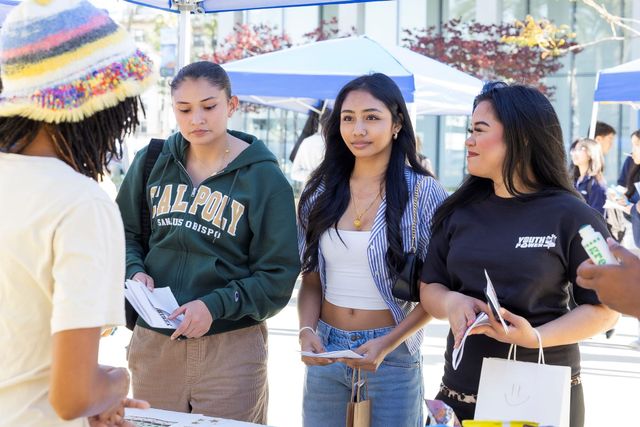 Three young people hold pamphlets while talking with another person underneath a tent typically found at fair