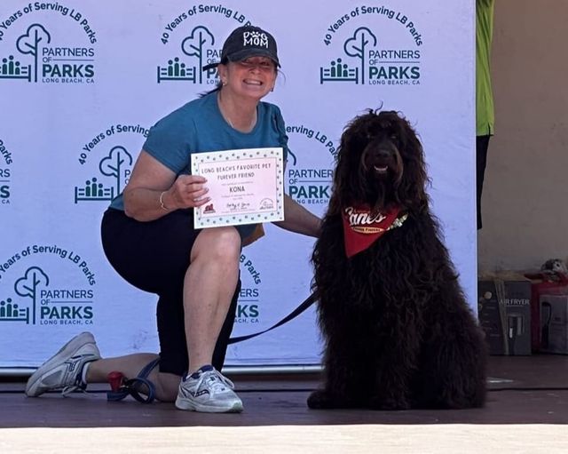 A woman holding an award certificate kneels on stage next to a large shaggy black dog that is wearing a red bandana.