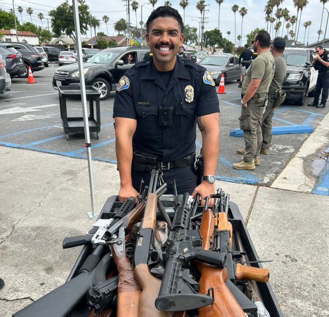 A smiling man in a police uniform stands next to a crate loaded with rifles in a parking lot surrounded by cars.
