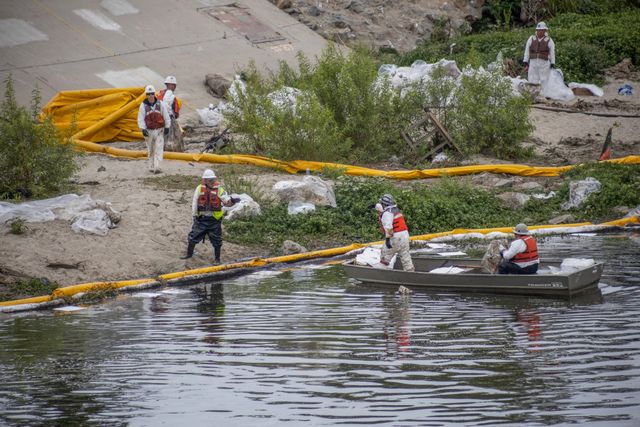 People dressed in white clothes with orange vests stand in a small gray boat on a gray river and on shore near a long yellow inflatable tube.