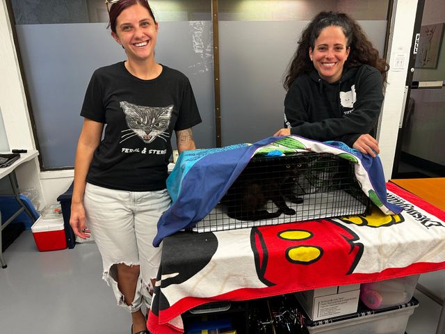 Two women smile while standing next to a partially concealed cage that holds a black cat.