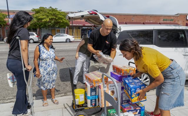 People stand on a sidewalk loading boxes of food and diapers into a white van.