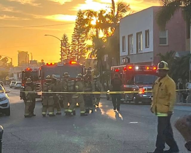 Firefighters stand around two fire trucks and an ambulance at sunset.