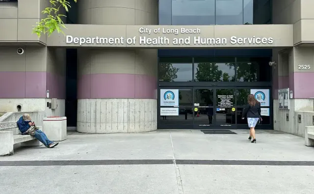 A woman walks into a building labeled "City of Long Beach Department of Health and Human Services" while a man sits on a bench nearby.