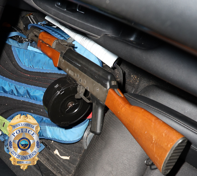 A rifle with a large round magazine rests against a vehicle se