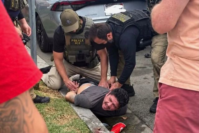 Men in body armor labeled "POLICE ICE" cuff and restrain a man lying in the gutter of a street while people look on.