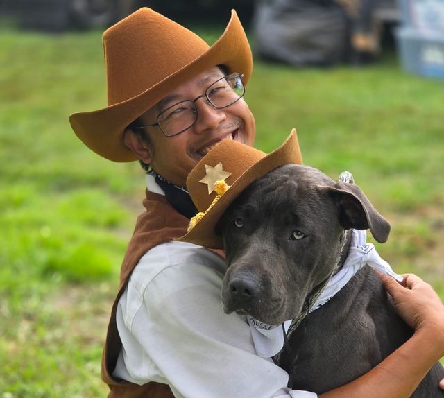 A man wearing a cowboy hat hugs a gray dog wearing a cowboy hat.