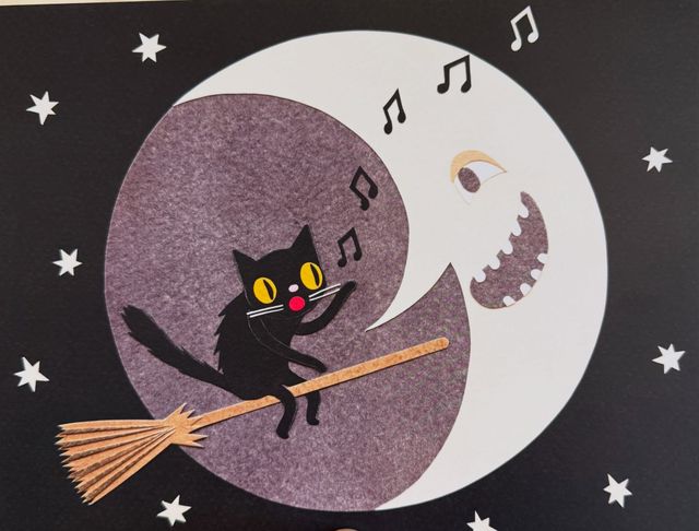 Paper-cut artwork showing a singing black cat on a broomstick in front of the moon at night with stars in the background.
