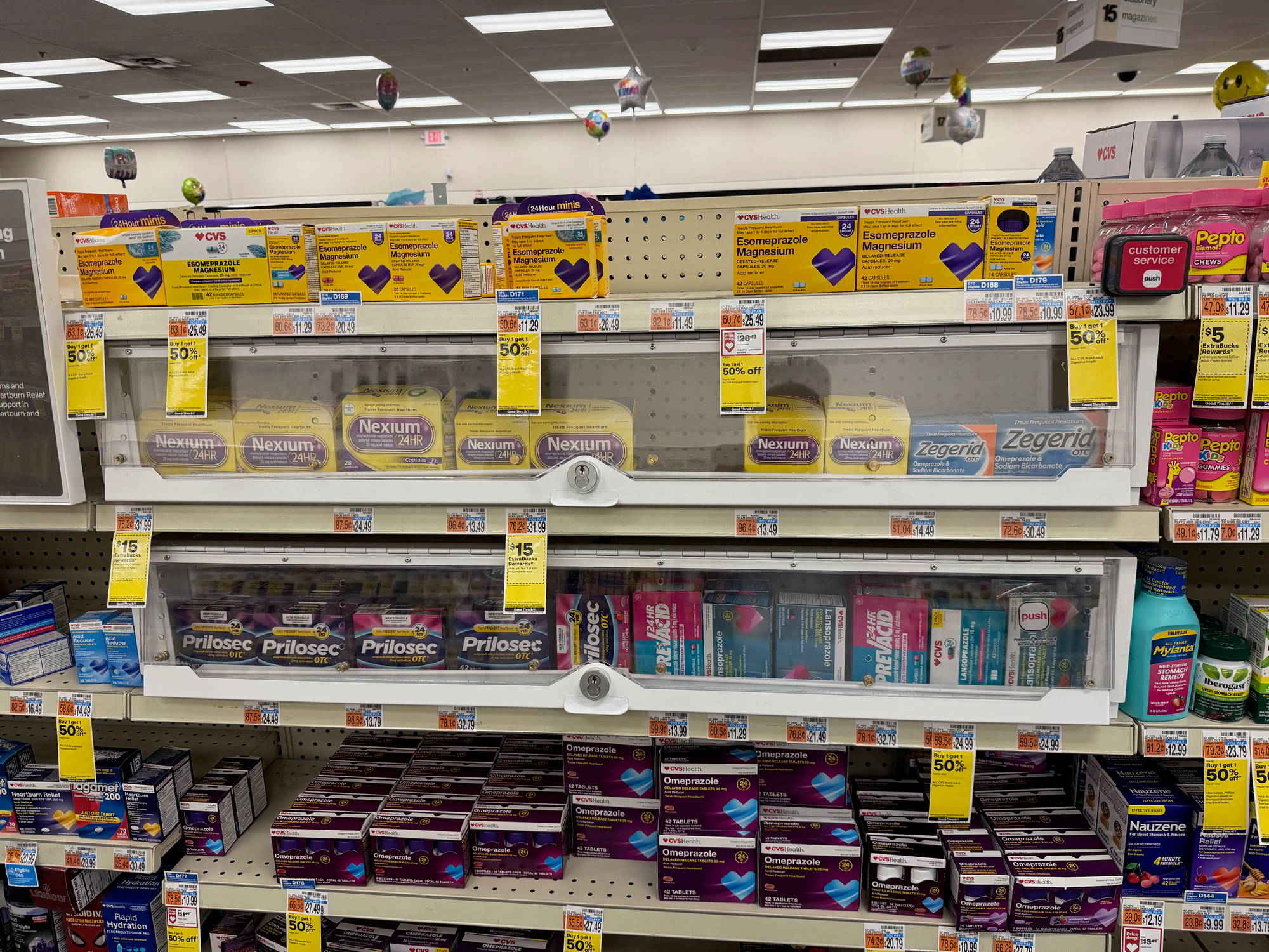A CVS shelf run of heartburn medication. Two long clear cases with keyed locks at their centers hold the brand names — Nexium 24HR and Zegerid OTC on the upper shelf, Prilosec OTC and Prevacid below. On the open shelves directly above and below the locked cases, CVS Health store-brand esomeprazole and omeprazole sit unlocked in stacks, their boxes reading "Compare to Prilosec OTC" and "Compare to the active ingredient in Nexium 24HR." A small red "customer service" button marked "push" is mounted at the far right end of the run, several feet from either lock, beside the Pepto-Bismol.