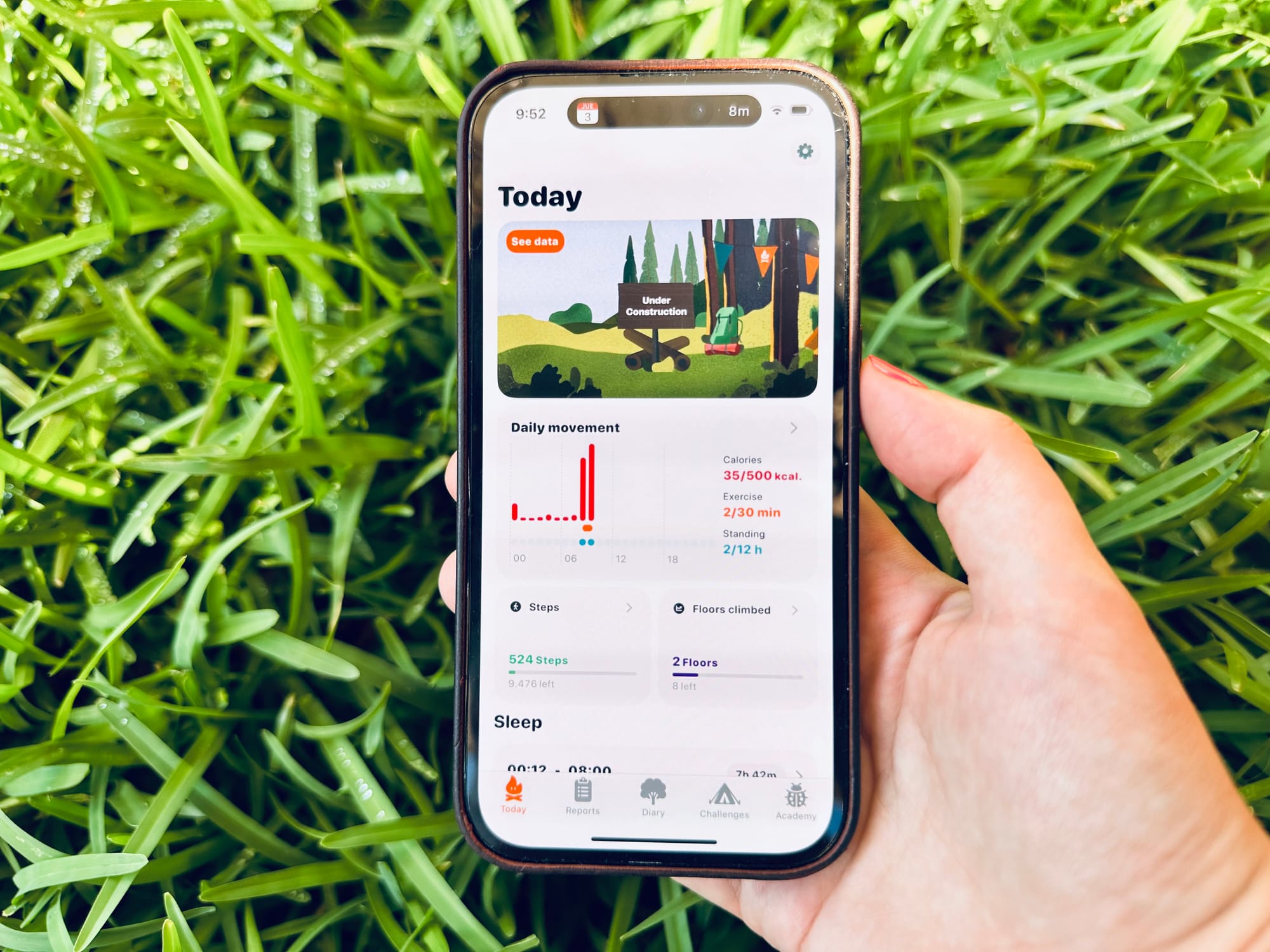 Hand holding a phone displaying one of the first versions of FitWoody, showing the “Today” dashboard screen with daily movement and a playful “under construction” illustration. The background is bright green grass.