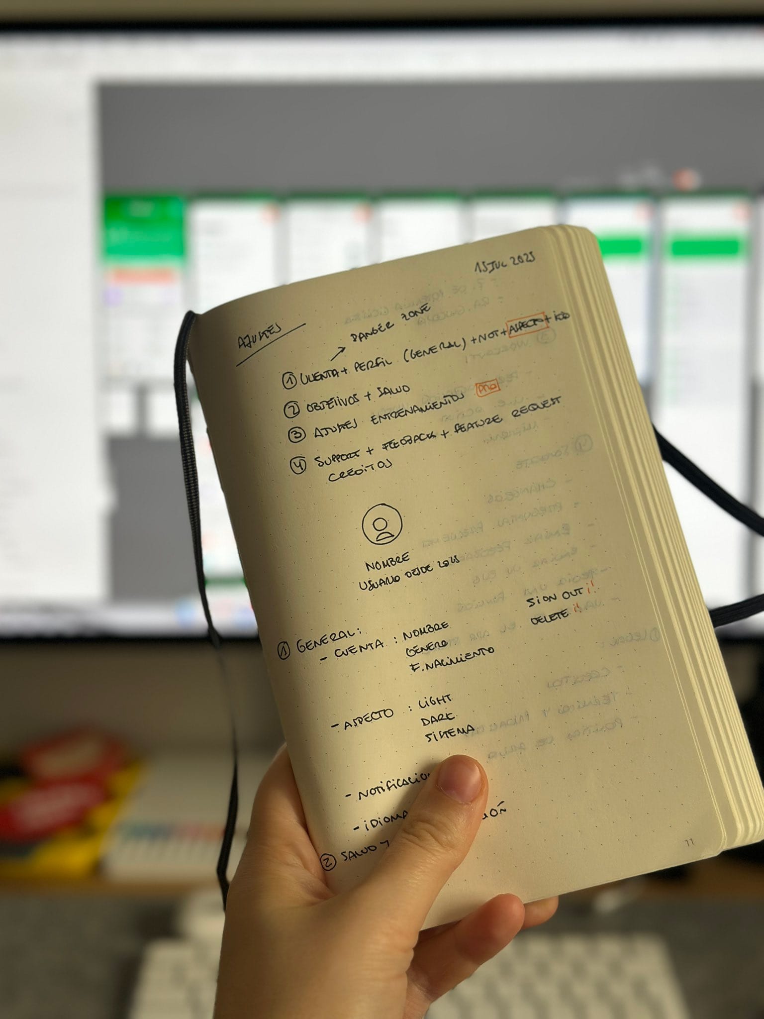 Hand holding an open notebook with handwritten notes and sketches for FitWoody’s early app features and user flow, dated July 2023. In the background, a blurred computer screen shows UI mockups in progress.