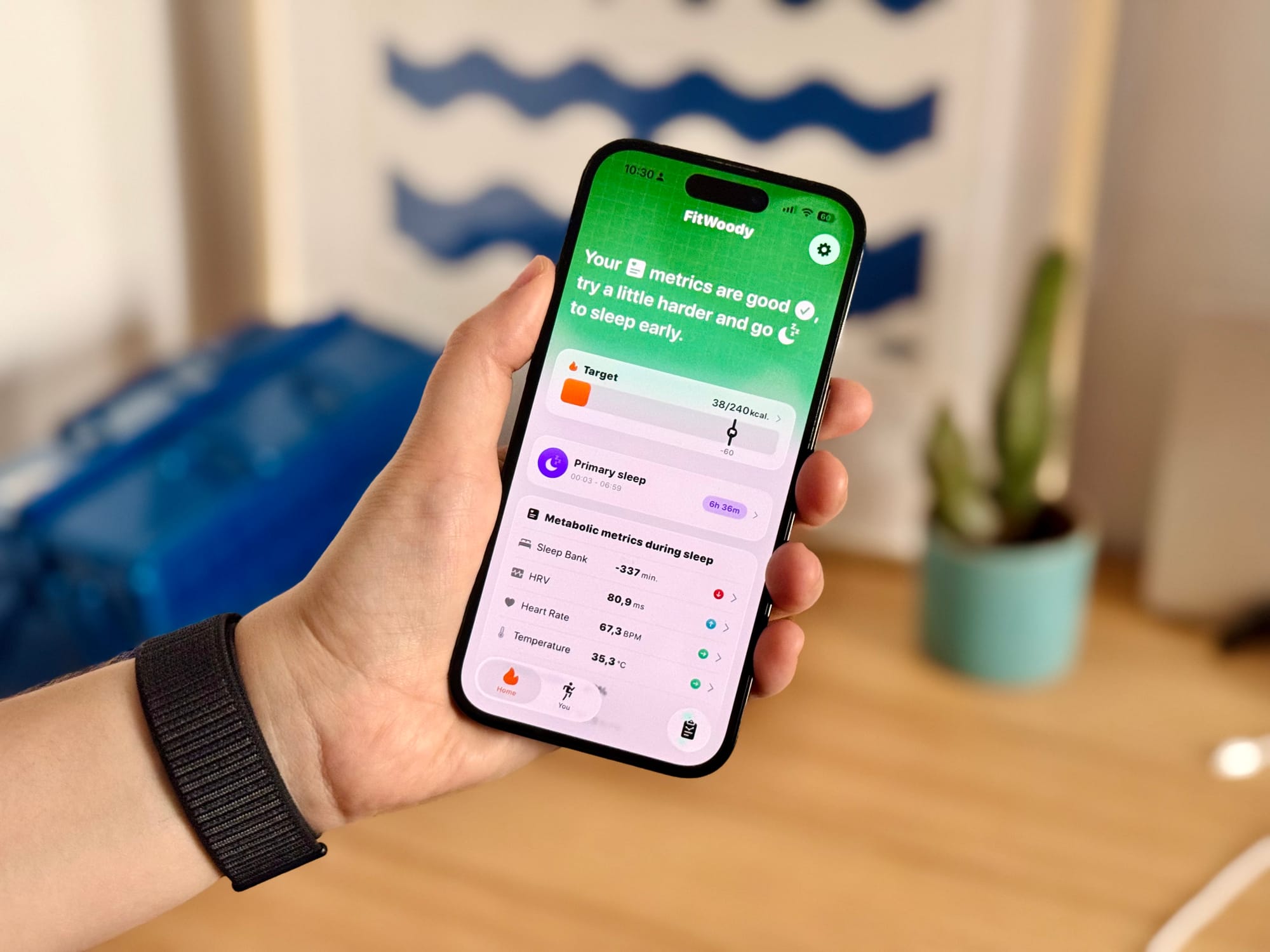 Hand holding a phone displaying FitWoody 2.0 with the redesigned dashboard, showing personalized health metrics, daily targets, and a sleep analysis summary. The background features a home or workspace with a blue object and a small potted plant.