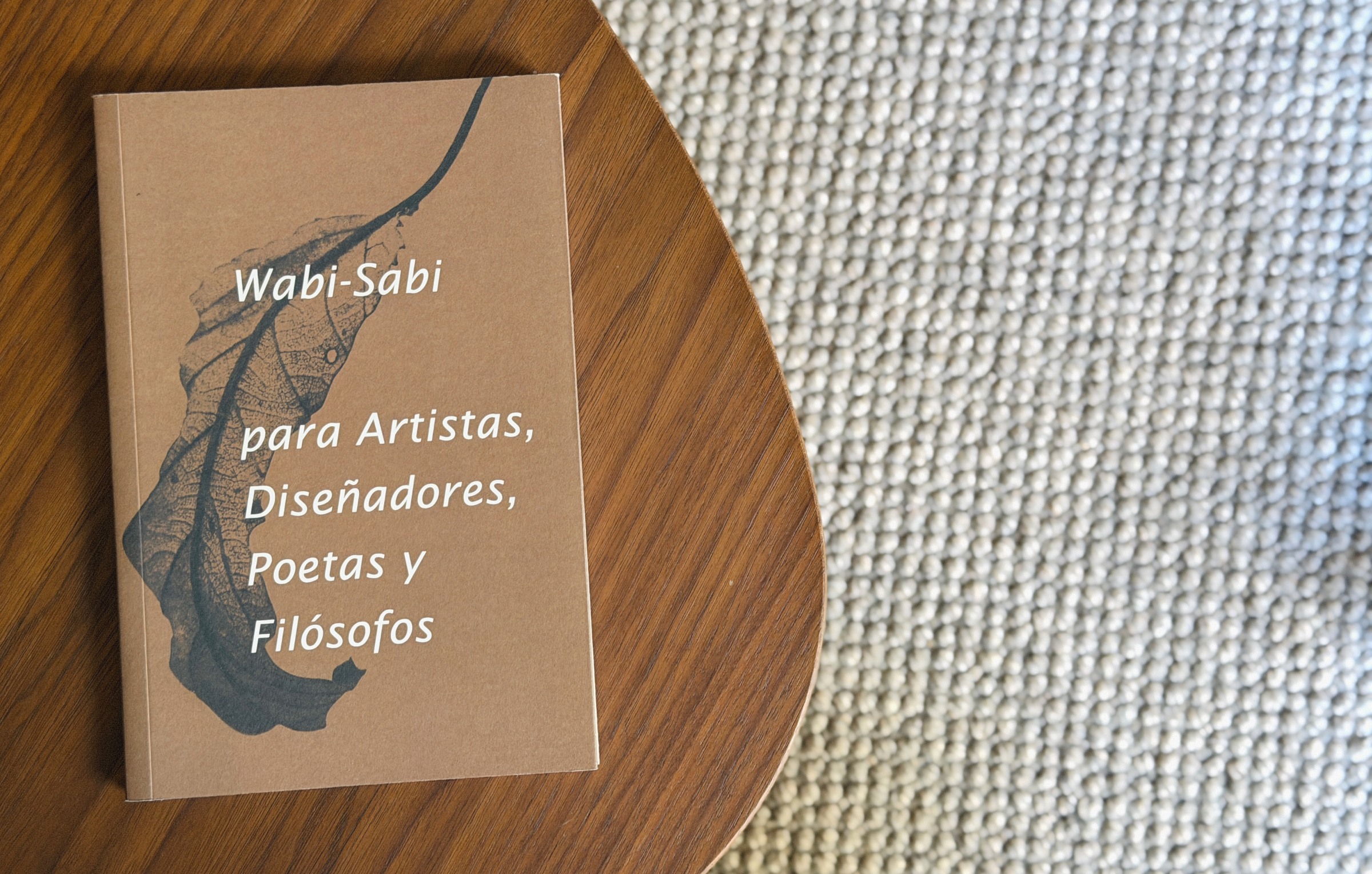 A book titled “Wabi-Sabi para Artistas, Diseñadores, Poetas y Filósofos” rests on a wooden table next to a textured beige carpet.