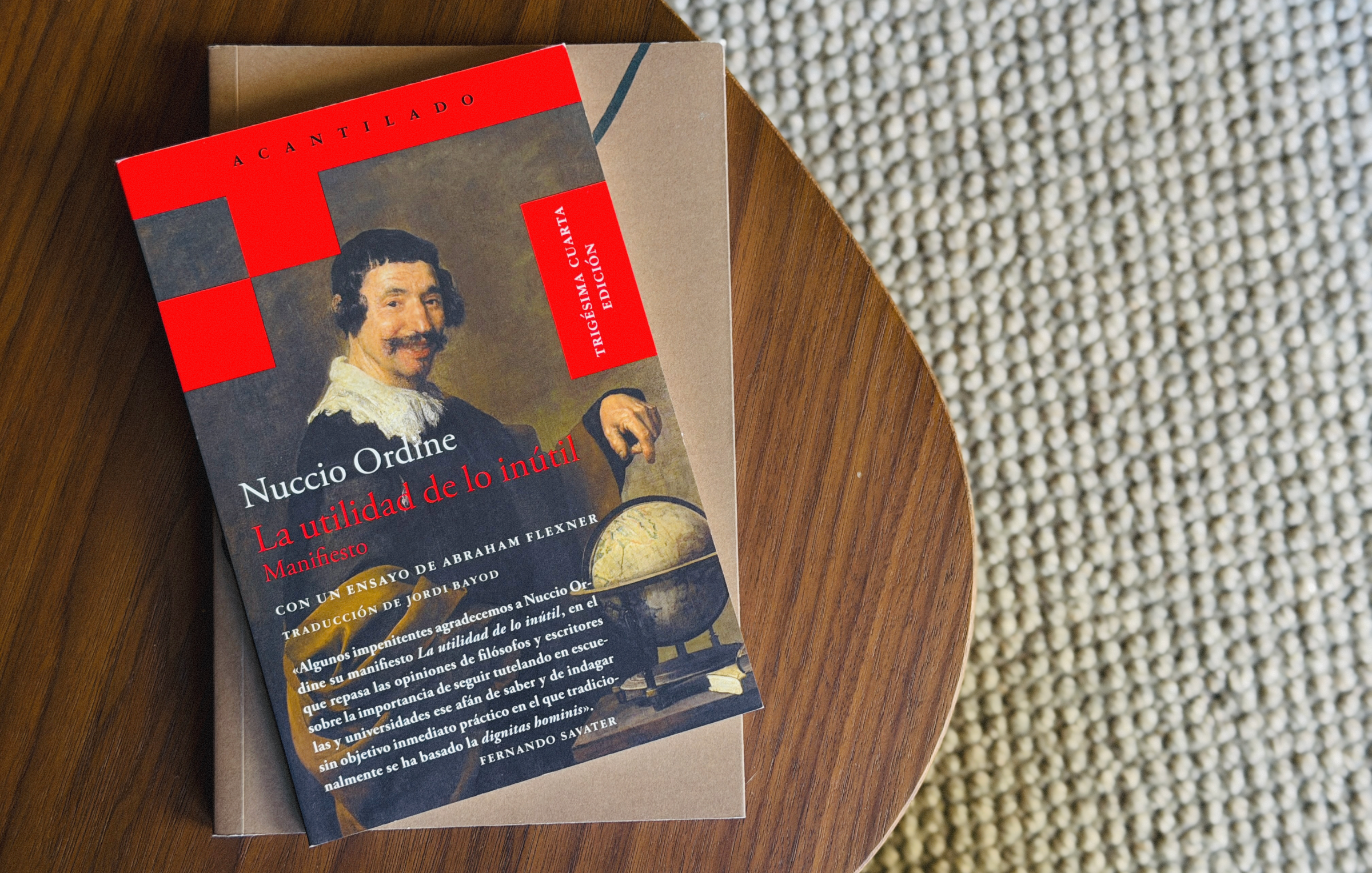 The book “La utilidad de lo inútil” by Nuccio Ordine rests on top of another book on a wooden table beside a textured beige carpet.