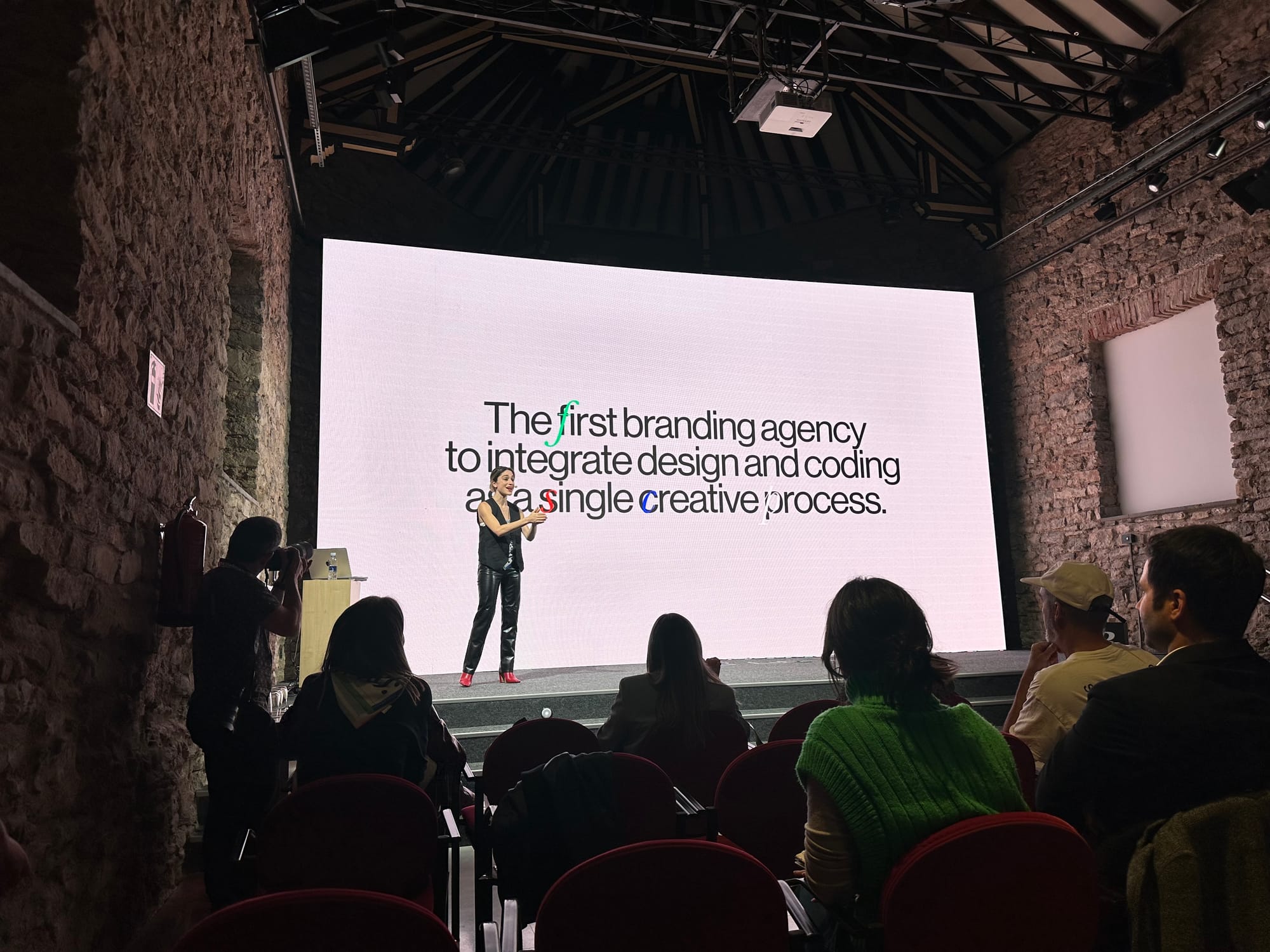 Talia Cotton on stage at Basquerville 2024, in front of a large screen that reads "The first branding agency to integrate design and coding as a single creative process", with the audience seated in the foreground of a stone-walled hall.