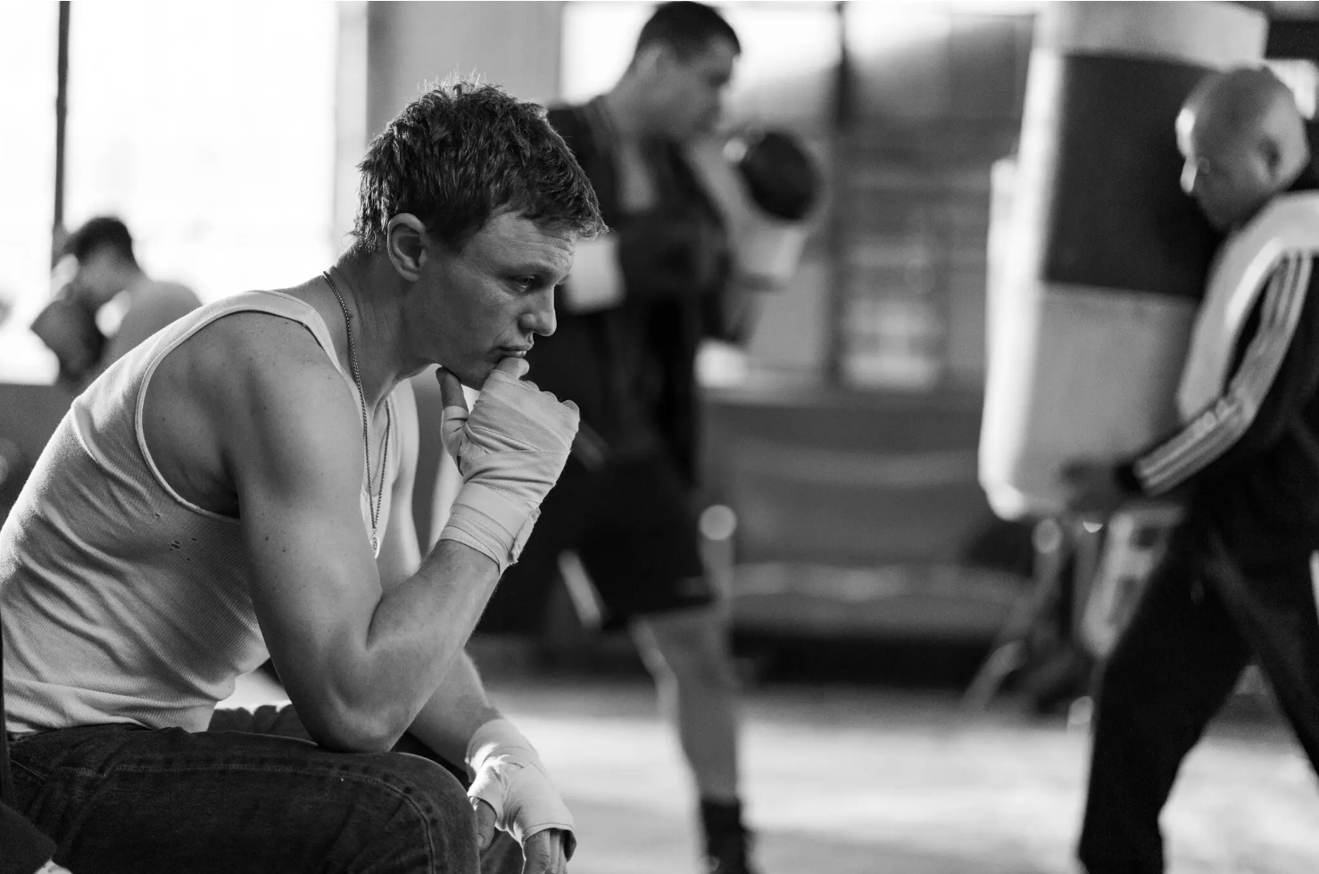 A boxer dressed in an a-frame undershirt and sweat pants, his hands taped, sits pensively in a boxing gym as other fighters work out in the background