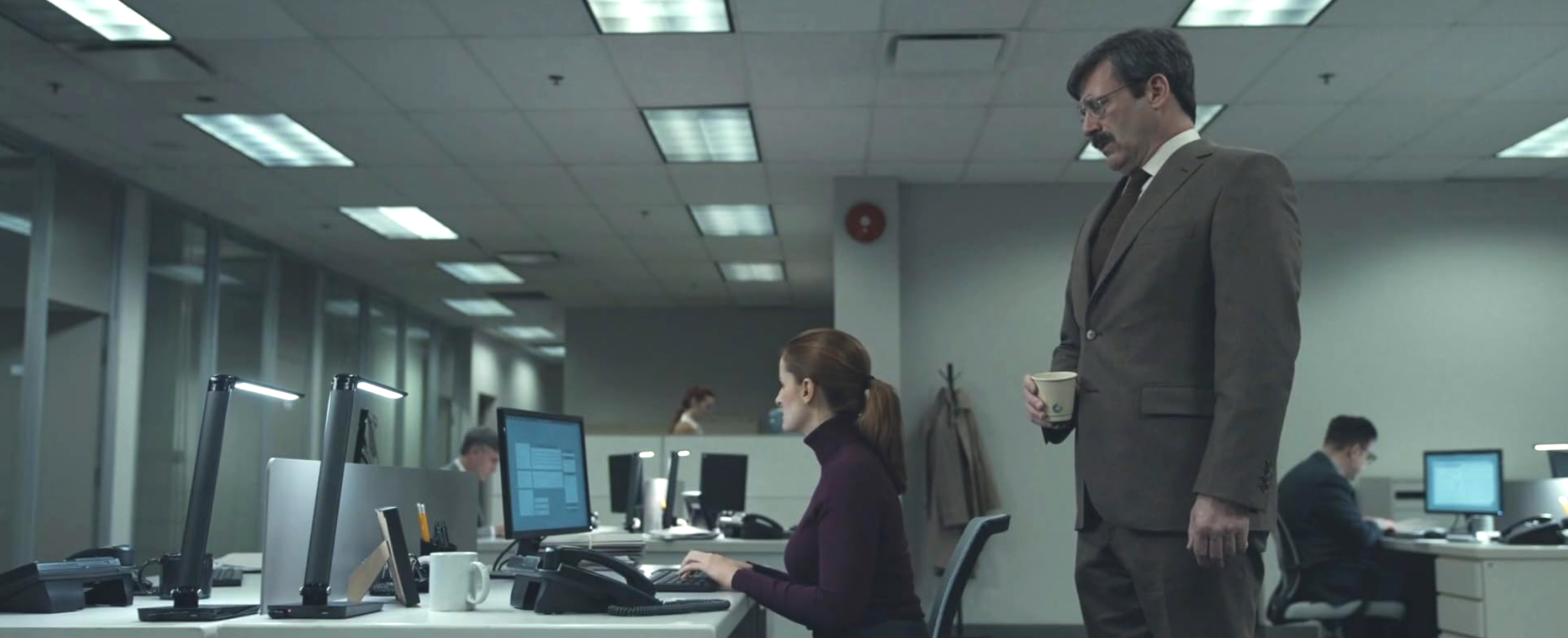 The actor Jon Hamm in a role as an office worker, standing and holding a cup of coffee while looking at a seated female coworker who does not acknowlege him, as other office workers toil in the background in a white-painted, florescent-lit corporate office environment.