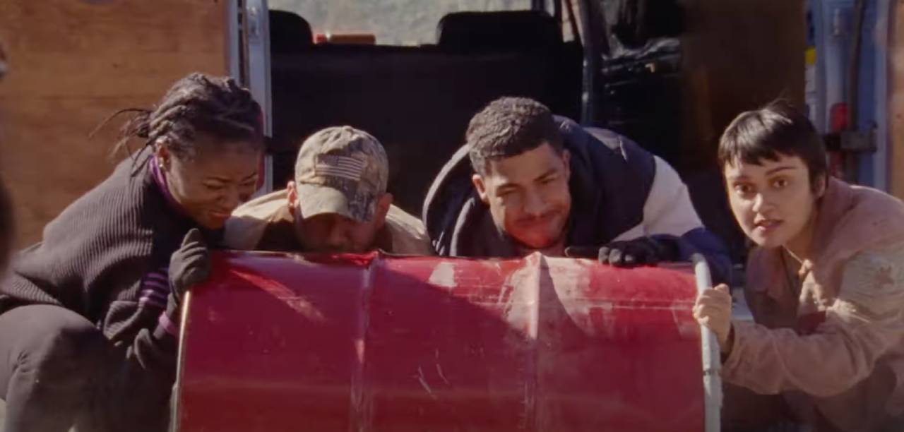 Four people grimace as they attempt to push a heavy red barrel.