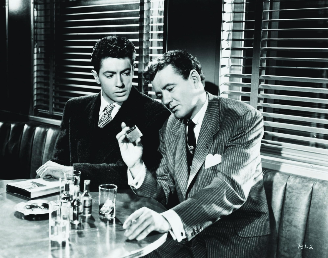 Two men dressed in early 1950s suits sit at a table in a passenger train lounge car. The man on the left is looking suspiciously at the other as the second man looks closely at a cigarette lighter. The image is in black and white.