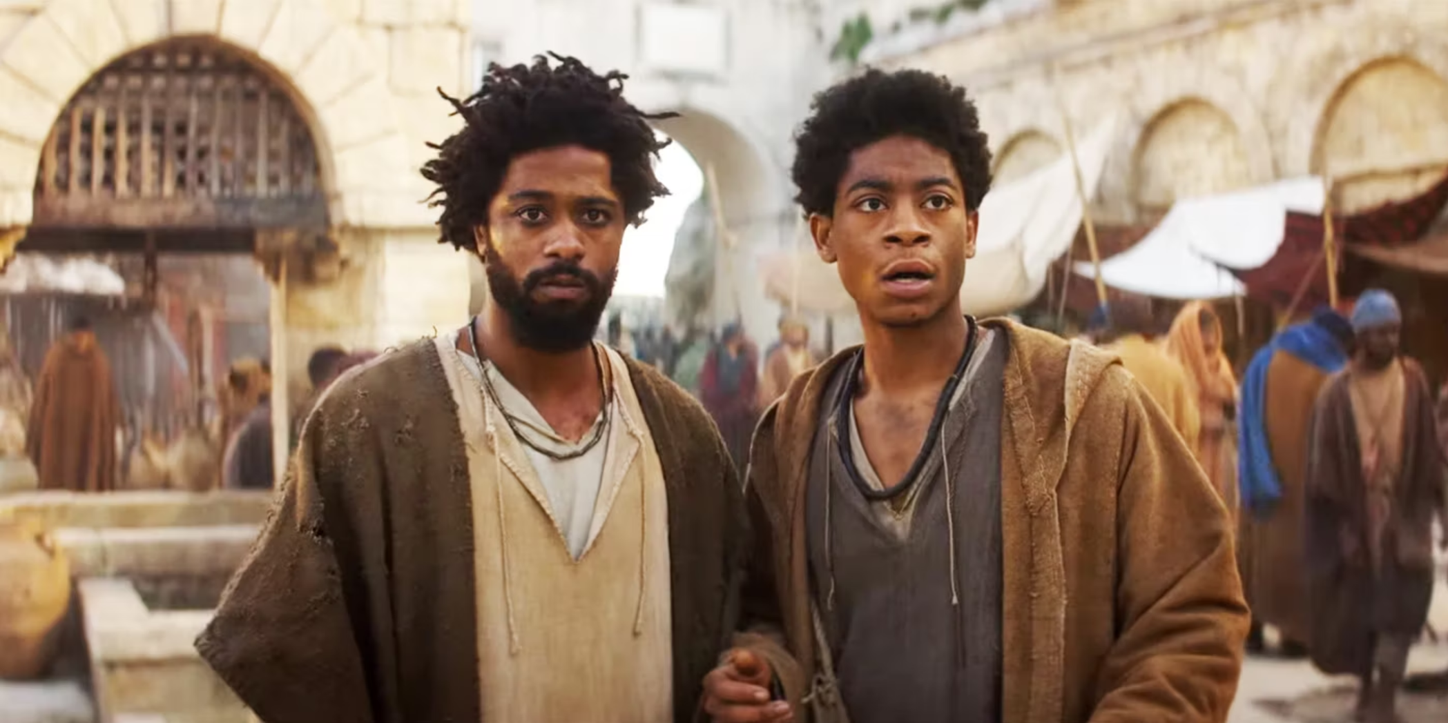 A film still from the movie “The Book of Clarence” shows two actors, LaKeith Stanfield and RK Cyler, dressed in brown and gray robes and tunics, with an ancient Mediterranean city scene in the background.