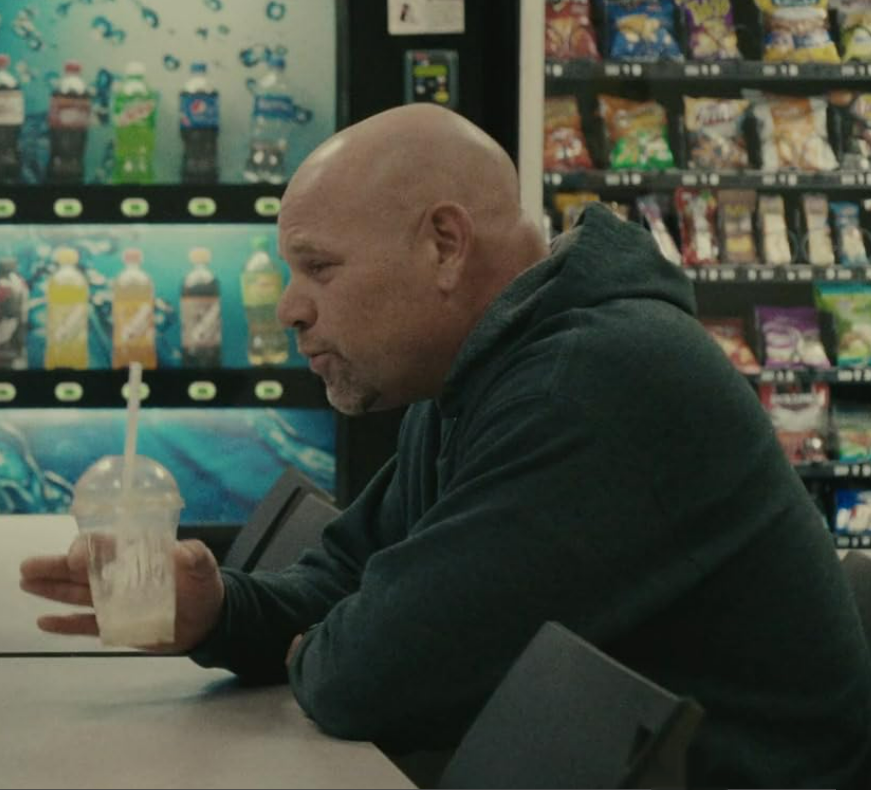 leDomenick Lombardozzi, a mille-aged, bald actor with a goatee, dressed in a green hoodie, leans on a breakroom table holding a mostly-consumed milkshake in a plastic cup, with vending machines in the background