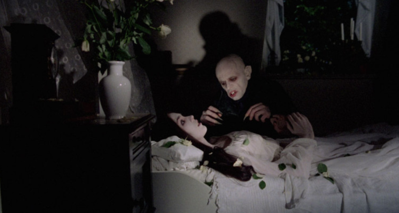 A frightening vampire extends his claws and leans over a woman lying in bed, her eyes open in terror. The woman is dressed in a white nightgown, while on a table by the bed sits a white vase with white roses