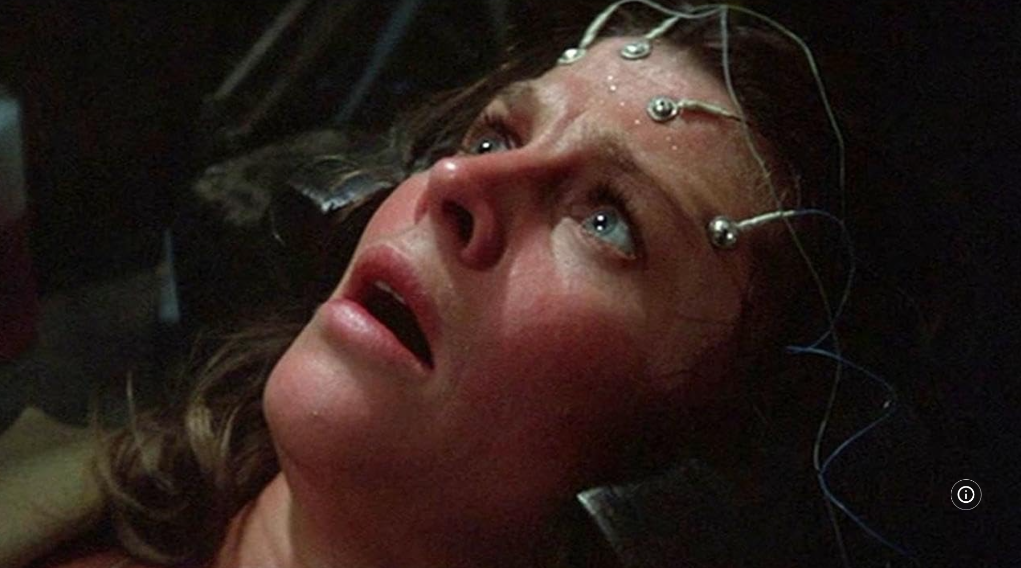 A woman (actress Julie Christie) wears an anguished expression and beads of sweat on her forhead, onto which are fasted several metal electrodes.