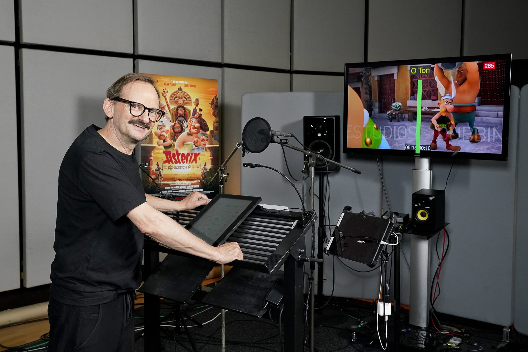 Milan Peschel steht lächelnd am Sprecherpult im Synchronstudio, hinten das Filmplakat
