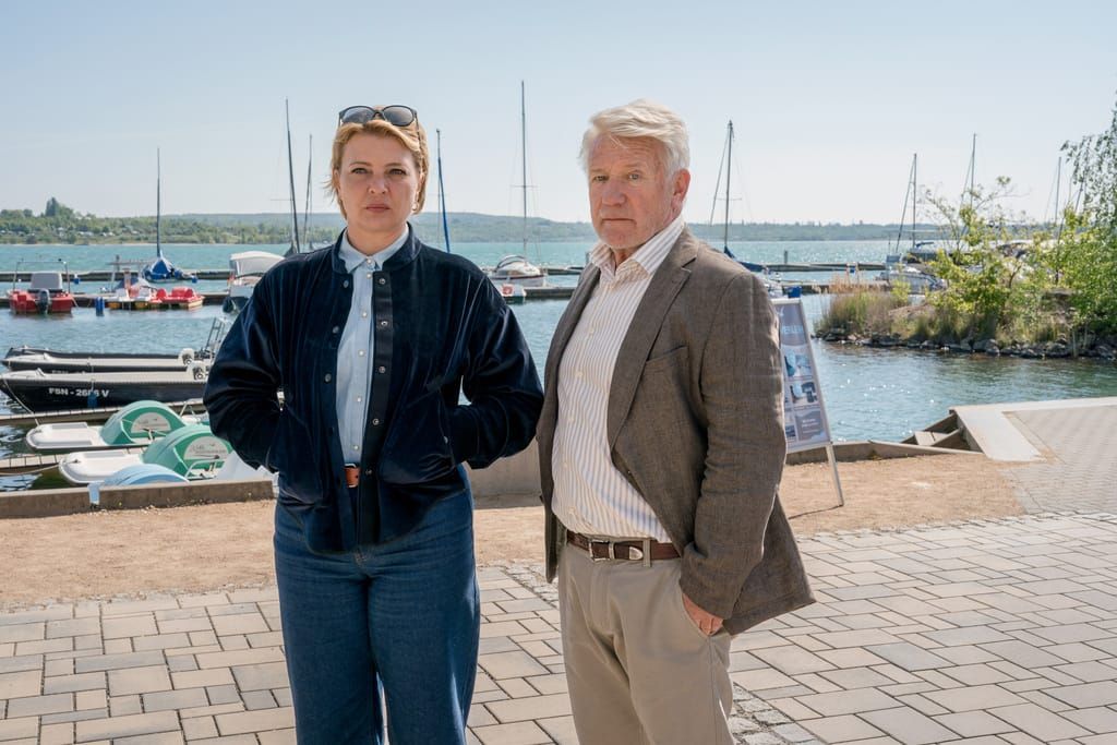 Jördis Triebel und Jörg Schüttauf stehen vor einem Hafen mit Segelbooten am See