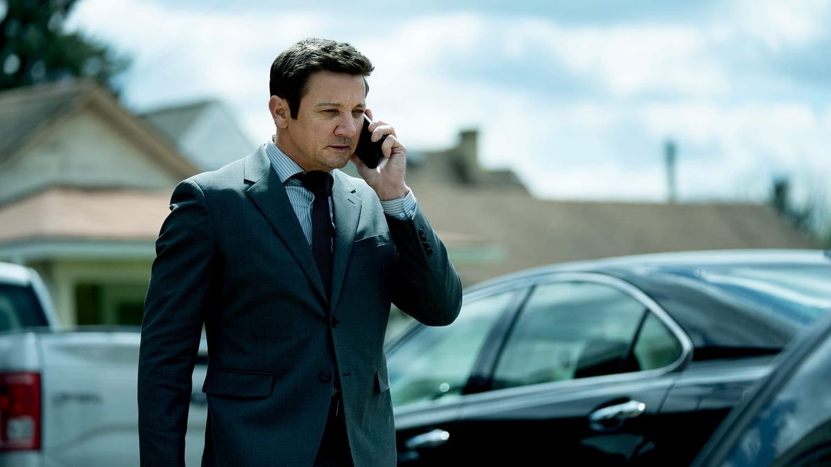 Jeremy Renner telefoniert im grauen Anzug zwischen geparkten Autos