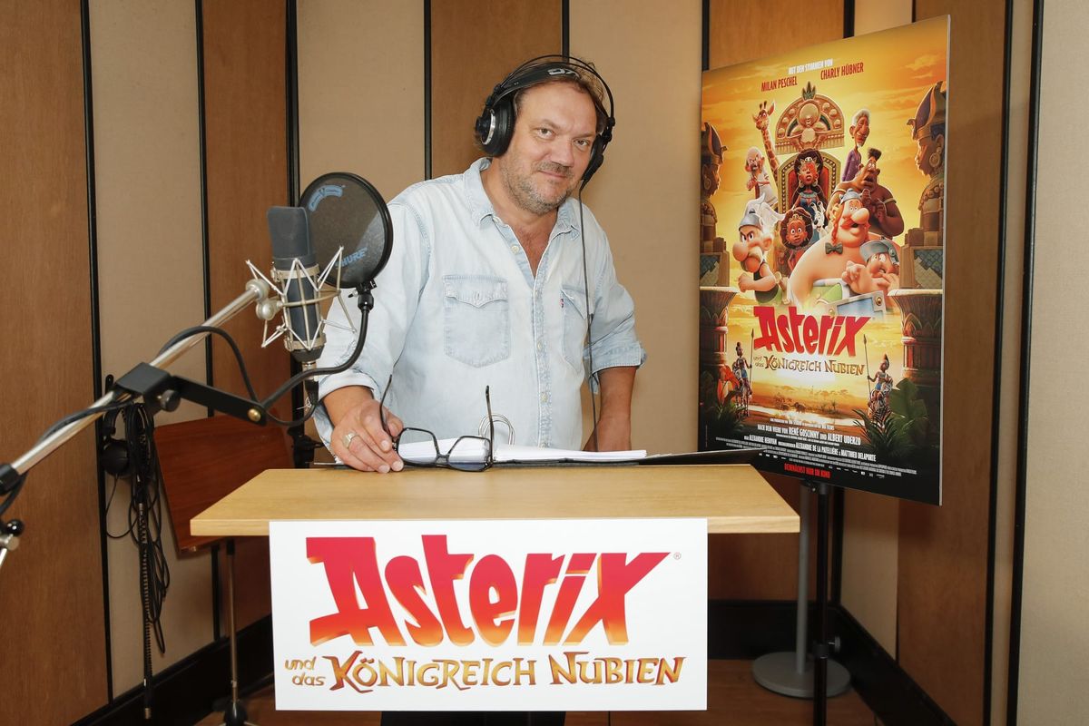 Charly Hübner mit Kopfhörern am Sprecherpult, daneben das Plakat zu „Asterix und das Königreich Nubien“