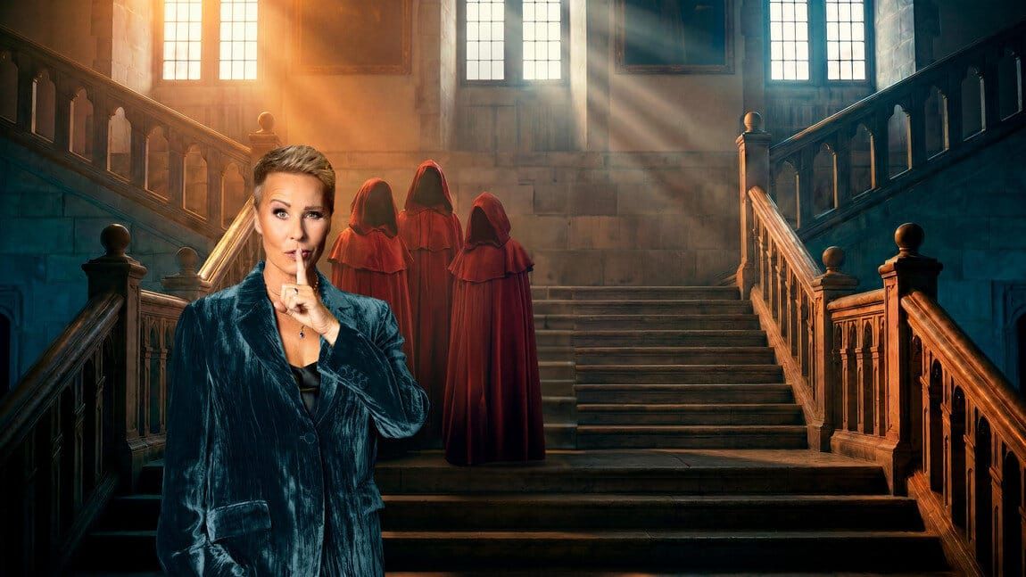 Sonja Zietlow mit Schweigegeste vor einer Schlosstreppe, dahinter drei Figuren in roten Kapuzenumhängen