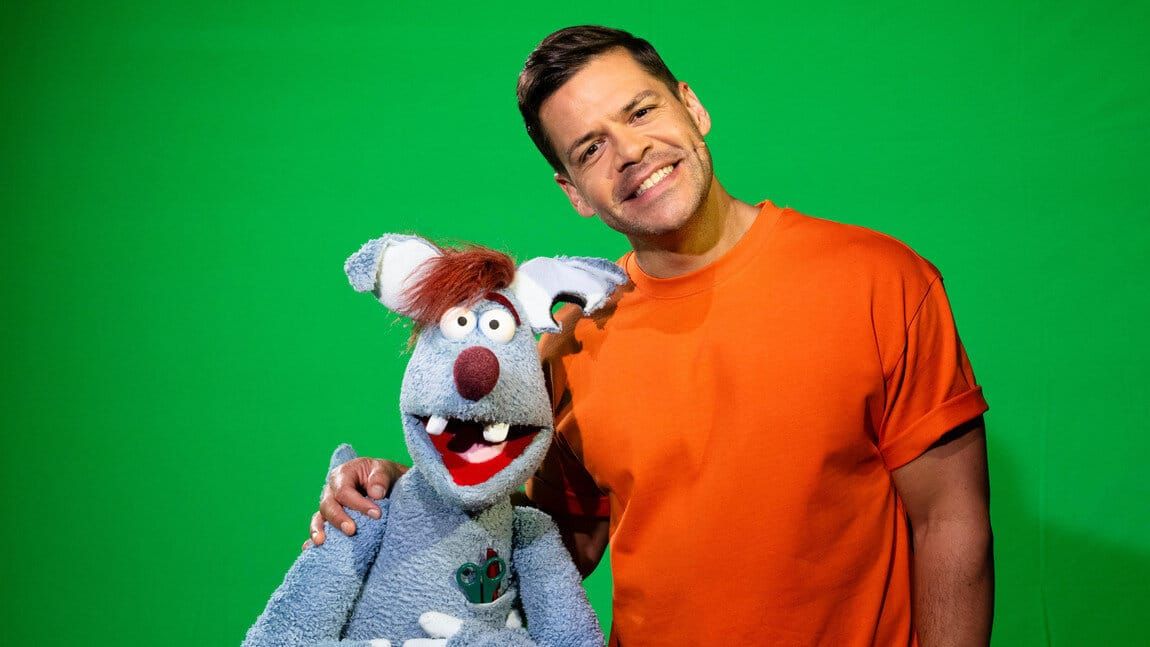 Moderator Simón Albers im orangen Shirt mit der Puppe Woozle vor Greenscreen