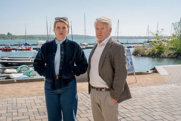 Jördis Triebel und Jörg Schüttauf stehen vor einem Hafen mit Segelbooten am See