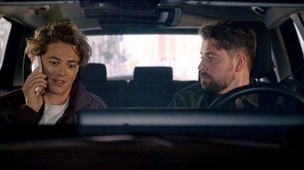 Benni (Julian Krenn) telefoniert im Auto, am Steuer sitzt sein Vater John Bachmann (Felix von Jascheroff)