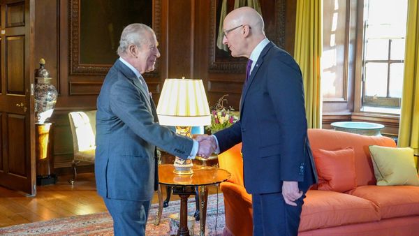König Charles III. im grauen Anzug schüttelt John Swinney in einem holzgetäfelten Salon des Holyrood Palace die Hand