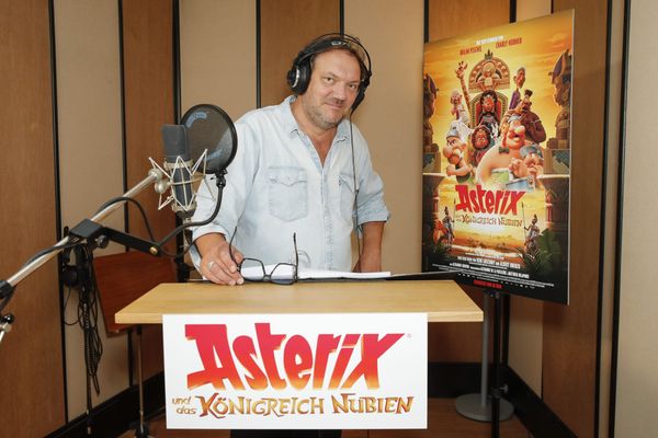Charly Hübner mit Kopfhörern am Sprecherpult, daneben das Plakat zu „Asterix und das Königreich Nubien“