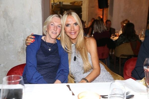 Zwei Frauen am gedeckten Tisch eines Abendessens, eine in Blau, eine im silbernen Kleid, Arm um die Schulter