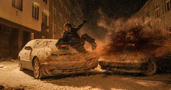 Eine Gestalt springt nachts in einer verschneiten Straße über ein Auto, umgeben von Funken und Staub