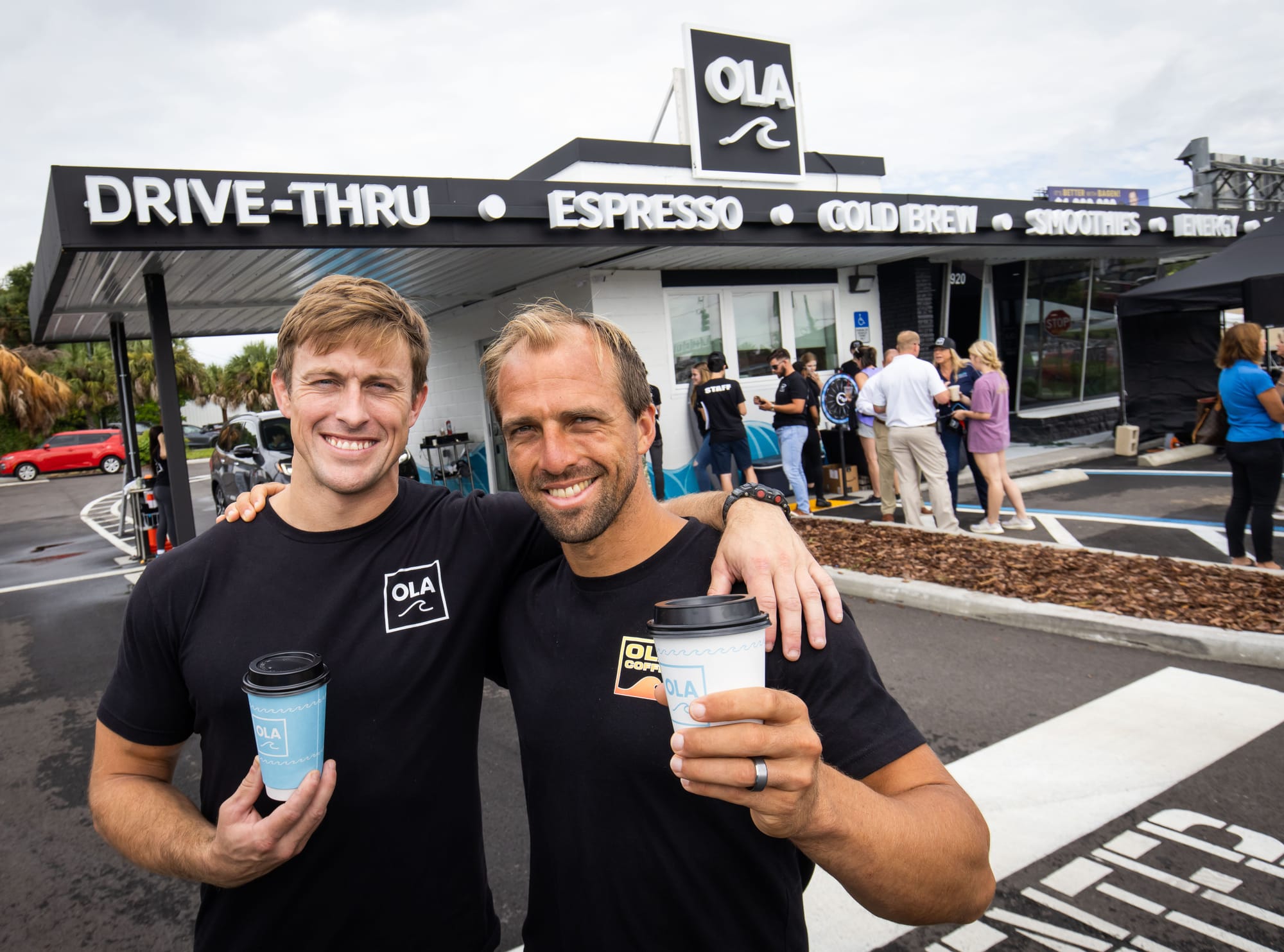 Corey Noel Scott, a white man with thinning dirty blonde hair and beard stubble, holds up a white cup of coffee with a light blue logo reading “OLA” and featuring an outline of a wave. A bold silver ring appears on his finger. Scott is wearing a black short-sleeve shirt with “OLA COFFEE” written on the side of the left breast pocket. Scott is smiling and putting his arm over another smiling white man with dirty blonde hair holding up an “OLA” branded coffee cup and black shirt. The two men are standing in front of the drive thru of a newly painted black and white small rectangle building with a single car drive thru awning. The white text along the top of the building reads “OLA” and “DRIVE-THRU, ESPESSO, COLD BREW.” About a dozen people are standing in line at the coffee shop’s walk up windows. A vehicle can be seen pulling into the drive-thru.
